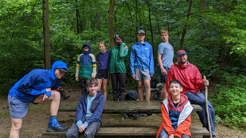 A group of people are gathered in a wooded area, likely on a hike or camping trip. They are standing and sitting around a wooden table. The group consists of both adults and children, dressed in casual outdoor clothing, including jackets and hats. The setting appears to be a forest, with trees visible in the background. The weather seems mild, as some people are wearing short sleeves. 